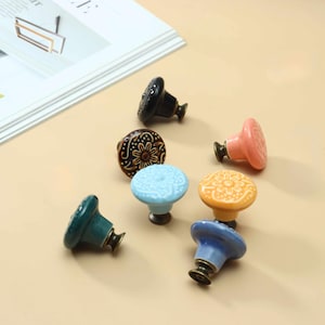 May include: A collection of seven ceramic cabinet knobs in various colors including black, dark brown with floral patterns, peach, light blue, yellow, dark teal, and medium blue, each featuring a metallic bronze-toned base and hardware.