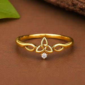 Könnte beinhalten: Ein Ring aus poliertem Gold mit einem zentralen Triquetra-Knoten-Design. Die Ringschiene ist glatt und kreisförmig, wobei das Knotenmotiv oben angebracht ist. Ein einzelner, kleiner, klarer, rund geschliffener Edelstein sitzt unter dem Knoten.
