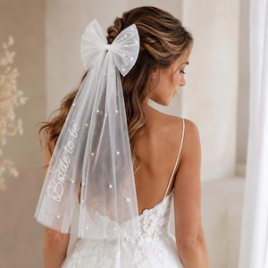 May include: A white tulle hair veil featuring a large bow at the top, scattered pearl embellishments, and the text "Bride to be" embroidered in white script. The sheer fabric drapes downward, creating a layered effect with delicate pearl accents.