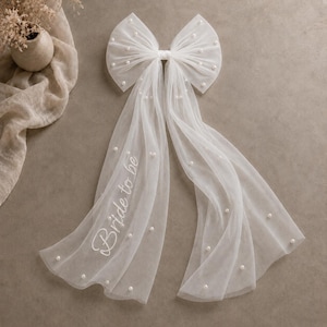 May include: A white tulle hair bow featuring scattered faux pearls and long, flowing ribbons. The left ribbon displays the text "Bride to be" in white cursive script. The sheer fabric creates a layered, delicate appearance with a soft texture.