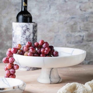 May include: A white marble pedestal bowl filled with a cluster of red grapes, positioned in front of a tall, cylindrical marble wine cooler containing a dark glass bottle. The marble features grey veining throughout the bowl and the cooler.