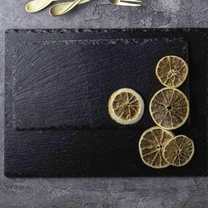 May include: Two rectangular black slate serving boards with rough, natural edges. The boards are stacked, and four dried, circular citrus slices are arranged on the right side of the top board. The surface of the slate has a textured, matte finish.