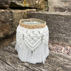 Peut inclure: Un bocal en verre transparent enveloppé d'une corde en macramé blanc avec un motif de losanges noués et une base à franges. Le bord supérieur du bocal est entouré d'une corde de jute tressée beige, offrant un contraste texturé.