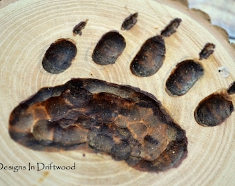 May include: A close-up of a wooden bear paw print, burned into a wood slice. The paw print is dark brown and the wood slice is light brown.