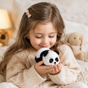 Op de afbeelding: Een klein, handgemaakt gehaakt pandabeertje met een rond wit lichaam, zwarte vlekken rond de ogen, zwarte oren en zwarte ledematen. De panda heeft een geborduurde zwarte neus en mond, en is voorzien van donkere veiligheidsoogjes.