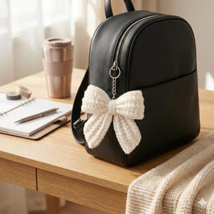 May include: A black leather backpack featuring a textured surface and a silver zip closure. Attached to the zip pull is a cream-coloured crocheted bow charm made of yarn. The backpack has a structured shape with a front pocket and shoulder straps.