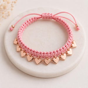 May include: A pink woven cord bracelet featuring a series of small, polished rose gold-toned heart charms dangling from the lower edge. The bracelet includes an adjustable sliding knot closure with two thin cord ends finished with small beads.