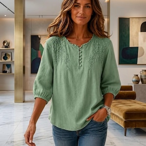 May include: A sage green blouse featuring three-quarter length sleeves with gathered cuffs, a round neckline with a ruffled edge, and a vertical row of small fabric-covered buttons. The upper chest area is decorated with delicate floral embroidery.