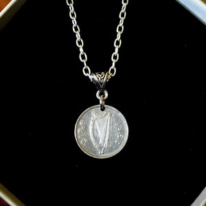 May include: Silver chain necklace with a silver coin pendant featuring the Irish harp and the year 1957. The coin is on a black background.