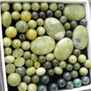 Op de afbeelding: Een verzameling groene en zwarte edelsteen kralen, sommige ovaalvormig en sommige rond, in een witte doos.
