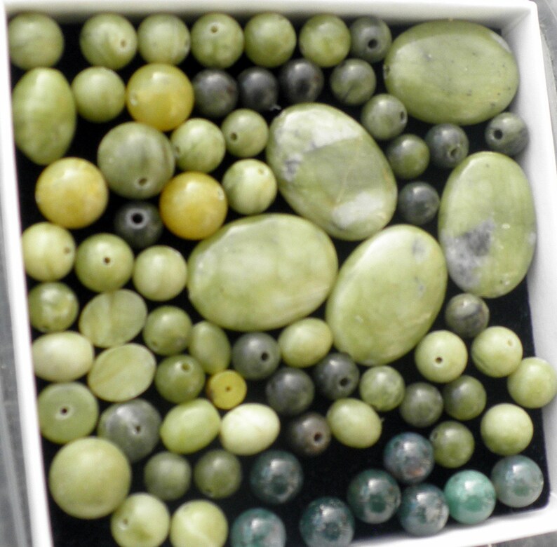 Op de afbeelding: Een verzameling groene en zwarte edelsteen kralen, sommige ovaalvormig en sommige rond, in een witte doos.