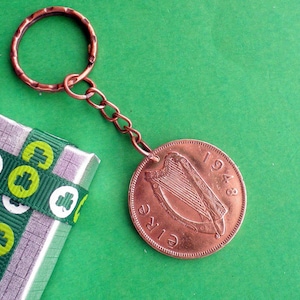 May include: A copper-colored keychain featuring a coin from 1948 with the word "EIRE" and a harp design. The keychain is attached to a copper ring and chain. A small gift box with green shamrock ribbon is in the background.