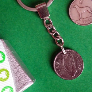 May include: A silver keychain with a chain and a 1943 Irish one penny coin attached. The coin features a harp design. A second coin with a greyhound is visible. The background is green.