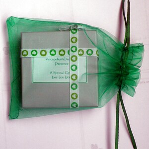 Op de afbeelding: Een klein, grijs cadeaudoosje met een groen-wit lint. Het lint heeft klavertjes en de tekst "Vintage Irish Originals Presents A Special Gift Just For You". Het doosje zit in een groene tule tas met een groen lint.
