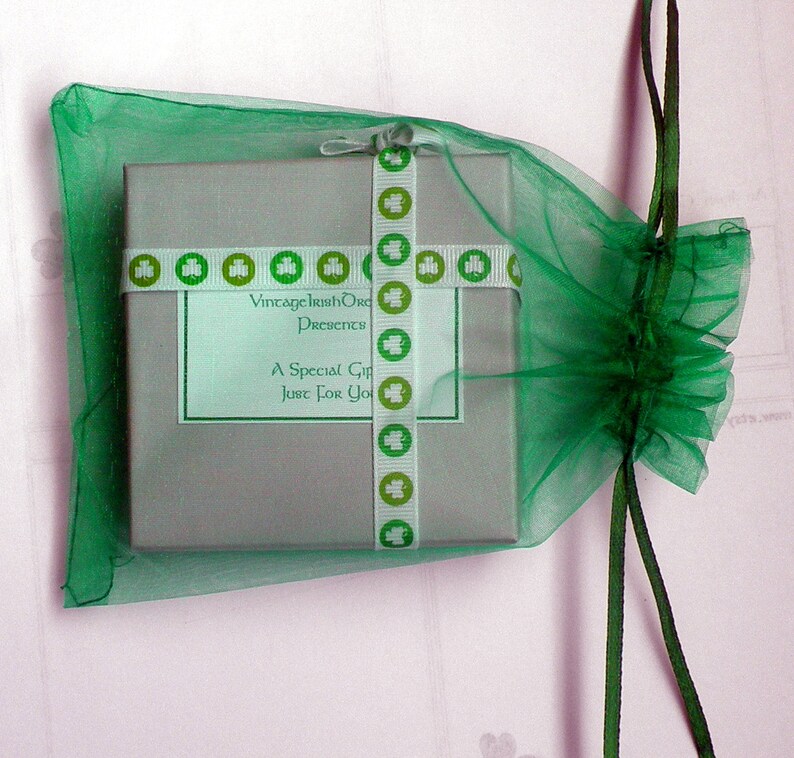Op de afbeelding: Een klein, grijs cadeaudoosje met een groen-wit lint. Het lint heeft klavertjes en de tekst "Vintage Irish Originals Presents A Special Gift Just For You". Het doosje zit in een groene tule tas met een groen lint.