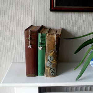 Op de afbeelding: Drie hardcover boeken met decoratieve bladwijzers. De bladwijzers zijn zilveren kettingen met groene kralen en een zilveren bedel. De boeken staan op een witte plank.