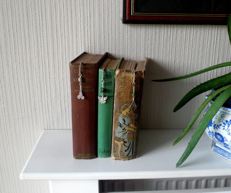 Op de afbeelding: Drie hardcover boeken met decoratieve bladwijzers. De bladwijzers zijn zilveren kettingen met groene kralen en een zilveren bedel. De boeken staan op een witte plank.
