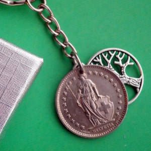 May include: Silver keychain with a Swiss coin featuring a woman holding a spear and shield with a cross. The coin has the word "Helvetia" inscribed on it. The keychain also includes a silver tree charm.