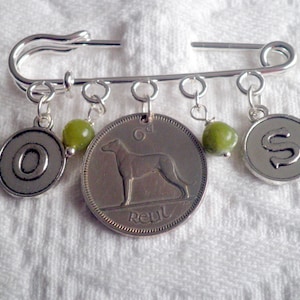 May include: A silver safety pin brooch with hanging charms. The charms include a coin with a dog image, a charm with the letter "O", a charm with the letter "S", and green beads. The brooch is on a white textured surface.