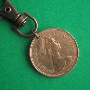 Op de afbeelding: Een koperkleurige sleutelhanger met een munt eraan bevestigd. De munt heeft een profiel van koningin Elizabeth II en de tekst "D.G. REG. F.D. 1967 ELIZABETH II".