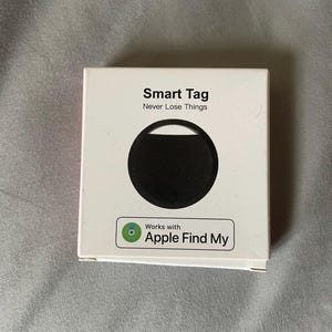 Könnte beinhalten: Eine quadratische weiße Produktverpackung mit einem schwarzen, kreisförmigen Smart Tag in der Mitte. Der Text lautet Smart Tag, Never Lose Things, und enthält ein grün-weißes Abzeichen mit der Aufschrift Works with Apple Find My.