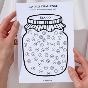 以下が含まれることがあります： $1,000とラベル付けされた瓶の黒い線画が描かれた白い紙の貯金チャレンジトラッカー。瓶の中には1、5、10、20、50と記されたコインを表す様々な円が含まれています。テキストにはSAVINGS CHALLENGE、Color in the coins to reach your goal、PAPER&COZYと書かれています。