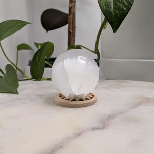 May include: A smooth, translucent white selenite sphere resting on a circular, light-coloured wooden stand with a geometric, star-like cutout pattern. The sphere has a small, rectangular white sticker with black text attached to its base.
