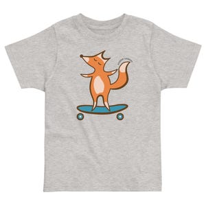 Może przedstawiać: Szary melanżowy t-shirt z krótkim rękawem z grafiką pomarańczowego lisa stojącego na niebieskiej deskorolce. Lis ma kremowy brzuch i zamknięte oczy. Napis "HUDSON LAUNDRY" jest wydrukowany w łuku nad ogonem lisa.