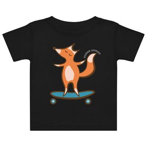 Może przedstawiać: Czarny t-shirt z krótkim rękawem z grafiką przedstawiającą pomarańczowego lisa z białym brzuchem stojącego na niebieskiej deskorolce. Napis HUDSON LAUNDRY jest wydrukowany w łuku nad ogonem lisa. Wzór posiada brązowe kontury.