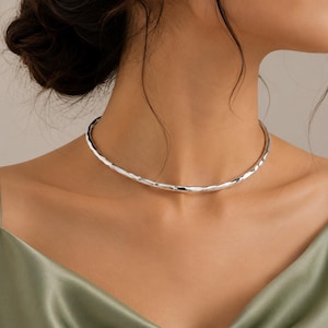 May include: A silver-toned choker necklace featuring a smooth, rounded band with a subtle, hammered, irregular texture along its surface. The necklace sits closely around the base of the neck, showcasing a reflective, metallic finish and a simple, circular design.