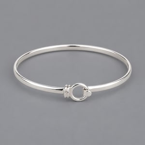 Puede incluir: Una pulsera rígida de tono plateado pulido con una banda circular lisa. El diseño incluye un elemento decorativo central que consiste en un pequeño anillo integrado, sujeto por una serie de tres estrías horizontales en un lado.
