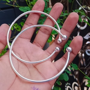 May include: Two silver-colored open-ended bangles, each featuring a smooth, thin circular band that terminates at both ends with a small, polished spherical ball. The bracelets have a bright, reflective metallic finish and a simple, minimalist design.
