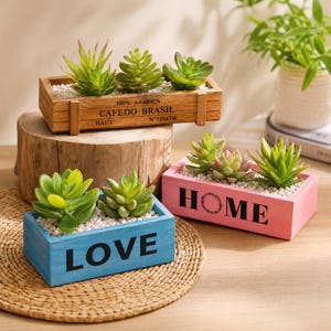 May include: Three rectangular wooden planters containing artificial green succulents and white pebbles. One planter is natural wood with "100% ARABICA CAFEDO BRASIL" text, one is painted pink with "HOME" text, and one is painted blue with "LOVE" text.