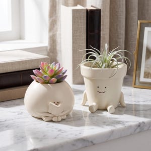 May include: Two cream-colored ceramic planters with simple facial features. One round planter depicts a figure reading a book and holds a multi-colored succulent. The other taller planter has small arms and legs and holds a spiky green air plant.