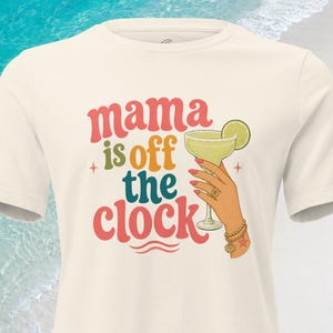 May include: A cream-colored short-sleeve t-shirt featuring the text "mama is off the clock" in red, orange, and teal. Next to the text is an illustration of a hand holding a margarita glass with a lime slice, accented by small star shapes.
