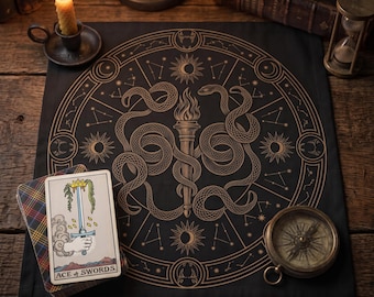 May include: A black square altar cloth featuring a gold-toned circular design with two snakes coiled around a central flaming torch, surrounded by celestial symbols and constellations, with a tarot card labeled Ace of Swords placed on top.