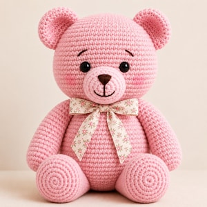 May include: A light pink crocheted teddy bear featuring a round head, small ears, and a stitched dark brown nose and mouth. The bear has black plastic eyes, rosy cheeks, and a cream-colored ribbon tied around its neck with a small floral pattern.