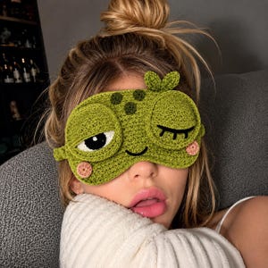 Puede incluir: Un antifaz de crochet verde con forma de cara de rana, con un ojo abierto que muestra una pupila negra y esclerótica blanca, un ojo cerrado guiñando, una pequeña sonrisa cosida, dos mejillas circulares rosadas y un pequeño detalle de hoja.