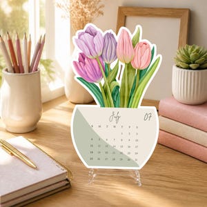 Può includere: Un calendario da scrivania per luglio con un'illustrazione ad acquerello di tulipani rosa e viola in un vaso. Il calendario mostra il mese di luglio, il numero 07 e una griglia di date dall'1 al 31 su uno sfondo beige e verde salvia.