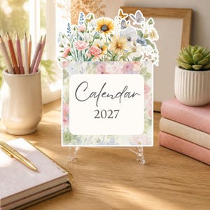 Op de afbeelding: Een vierkante kalender voor 2027 met een bloemenillustratie van zonnebloemen, madeliefjes en vlinders aan de bovenzijde. Het witte middenpaneel toont de tekst Calendar 2027 in een donker script en schreeflettertype, omringd door een bloemenrand.