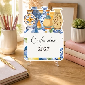 May include: A 2027 calendar featuring a decorative top border of various illustrated ceramic vases in blue, yellow, and tan hues. The central white rectangular area displays the text Calendar 2027 in a dark gray script and serif font.
