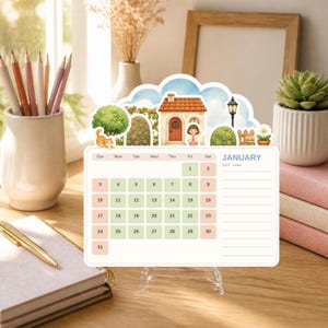 May include: A desktop calendar for January 2027 featuring a die-cut illustration of a house, a cat, and a person. The calendar grid displays dates in light green and pink squares, with a lined notes section on the right side. The design includes a blue sky background with clouds, a street lamp, and greenery.