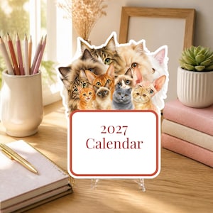 May include: A 2027 calendar featuring a collage of various cat breeds including tabby, Siamese, and hairless cats. The calendar has a white rectangular base with red text that reads 2027 Calendar, displayed on a clear stand on a wooden desk.