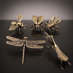 May include: A collection of five brass-toned metal insect figurines including a bee, a beetle, a grasshopper, a dragonfly, and a praying mantis. Each insect features detailed textured wings, segmented bodies, and articulated legs in a metallic finish.