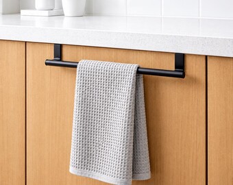 Pode incluir: Um toalheiro de metal preto fosco projetado para pendurar na porta de um armário, com um sistema de suporte retangular. Uma toalha de cozinha cinza-claro com um padrão de tecido waffle texturizado está pendurada na barra horizontal.