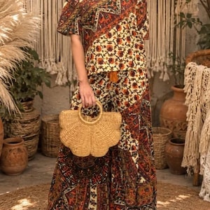 May include: A two-piece outfit consisting of a short-sleeved V-neck top and wide-leg pants, both featuring a multi-colored patchwork pattern in shades of brown, orange, and cream. A tan woven straw handbag with a fan-shaped design is held.