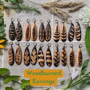 May include: A collection of 16 pairs of woodburned earrings. Each pair features a unique design, including feathers, leaves, and geometric patterns. The earrings are made of natural wood and have a rustic, handmade aesthetic.