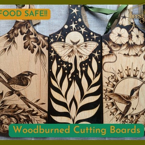 May include: Three woodburned cutting boards with different designs. The first board features a wren with a nest of eggs, the second board features a moth with a moon and stars, and the third board features a hummingbird with a sun design. The boards are all made of light wood and have a natural finish. The text "FOOD SAFE!" and "Woodburned Cutting Boards" is visible on the image.