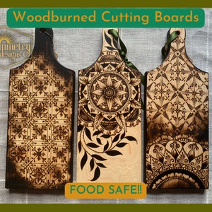 May include: Three woodburned cutting boards with different designs. The boards are all food safe and have a handle. The designs include a geometric pattern, a mandala, and a floral design.