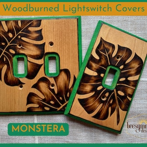 May include: Two woodburned light switch covers with a monstera leaf design. The covers are painted with a green border and have a natural wood finish. The text "MONSTERA" is printed below the covers.